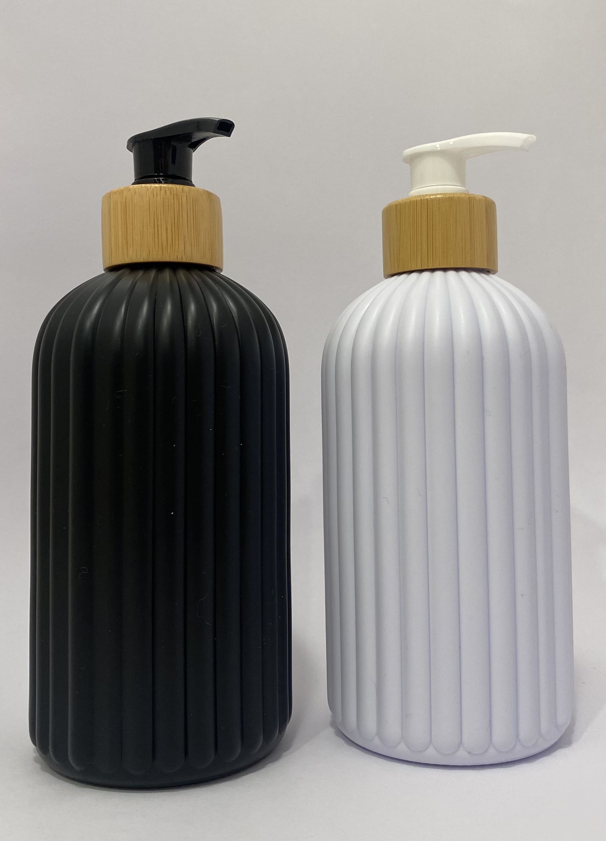 Two ribbed pump bottles on a light gray background, one black with a bamboo-look gold cap and black pump, and one white with a bamboo-look gold cap and white pump.